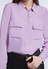 Blusa lilla chiaro con colletto a bottoni, due tasche sul petto e una superficie liscia e leggermente lucida, indossata con pantaloni neri.