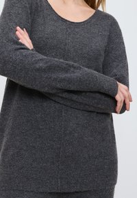 Frau, die einen dunkelgrauen Strickpullover mit langen Ärmeln trägt, die Arme verschränkt, vor einem schlichten hellen Hintergrund.
