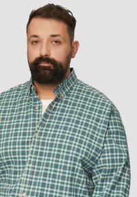 Man met kort donker haar en baard, gekleed in een teal, crème en rode geruite overhemden met een witte onderhemd, tegen een effen achtergrond.