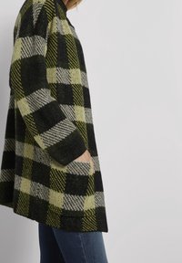 Dlouhý svetr v černé, šedé a limetkově zelené kostkované vzoru s texturovanou tkaninou. Má dlouhé rukávy a boční kapsy.