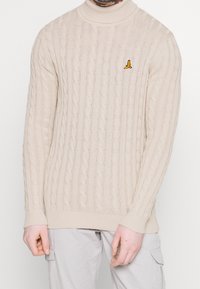 Beige Kabelstrickpullover mit einem enganliegenden Rollkragen. Besitzt gerippte Bündchen und Saum sowie einen kleinen gelben Vogel, der auf der Brust gestickt ist.