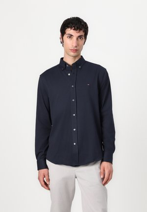 Cămașă navy cu guler, cu nasturi albi și un mic logo pe piept. Material din bumbac neted, mâneci lungi.