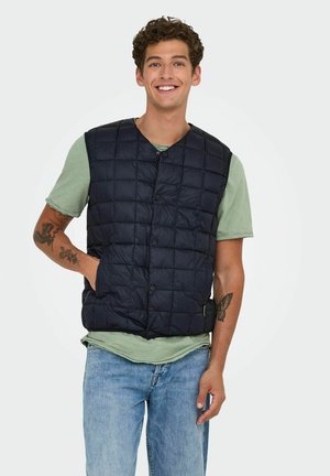 Gilet rembourré matelassé bleu marine avec une fermeture à boutons et deux poches latérales, porté sur un t-shirt vert clair, accompagné de jeans bleu clair.