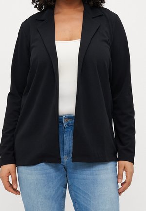 Vrouw in een zwarte open-front blazer over een witte top en lichtblauwe spijkerbroek, weergegeven van schouders tot heupen tegen een effen achtergrond.