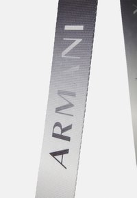 Szürke átmenetes szövet szalag, rajta vastag fekete betűkkel az "ARMANI" felirattal, texturált felülettel és sima szélekkel.