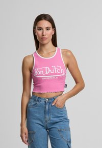 Jauna sieviete ģērbjas rozā īsajā topā ar uzrakstu "Von Dutch Originals" un augsto jostasvietu zilos džinsos, stāvot pret vienkāršu pelēku fona.