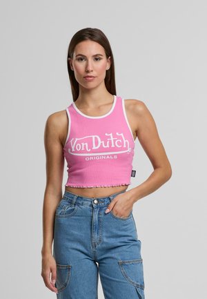 Noor naine kannab roosa lühikeste varrukatega tank-topp, millel on kiri "Von Dutch Originals", ja kõrge vöökohtadega siniseid teksaseid, seistes ühetoalise halli tausta vastu.
