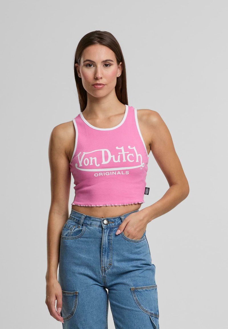 Jauna sieviete ģērbjas rozā īsajā topā ar uzrakstu "Von Dutch Originals" un augsto jostasvietu zilos džinsos, stāvot pret vienkāršu pelēku fona.