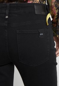 Jeans de mezclilla negra con una textura suave, que presentan un bolsillo trasero, una etiqueta de cuero y un bucle de acento amarillo. Detalles de costura sutiles presentes.