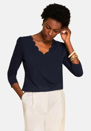 Frau mit kurzen lockigen Haaren, die ein marineblaues, gewelltes V-Ausschnitt-Top, weiße Hosen und goldene sowie cremefarbene Armreifen trägt, schaut nach unten und hält die Hand nahe am Ohr.