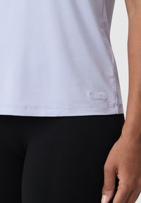 Detailní záběr na osobu v jasně fialovém tričku a černých kalhotách, s výšivkou "LES LUNES" poblíž lemu.