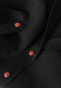 Tissu noir avec trois boutons ronds rouges encadrés d'or, disposés verticalement sur un vêtement plié.