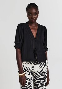 Blusa nera a trama con scollo a V profondo, maniche a sbuffo corte e dettaglio con fiocco in vita. Abbinata a pantaloni a righe.