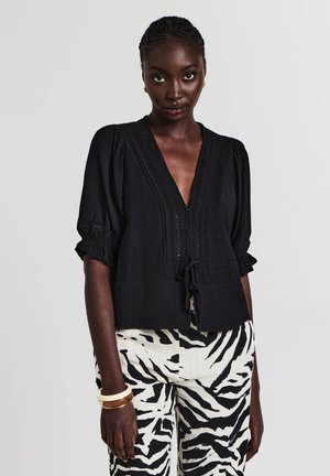 Schwarz strukturierte Bluse mit tiefem V-Ausschnitt, kurzen Puffärmeln und Bindedetail in der Taille. Kombiniert mit gestreiften Hosen.