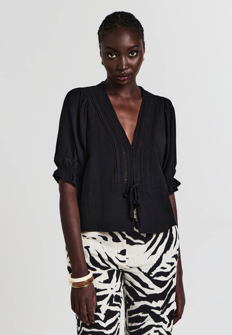 Blusa nera a trama con scollo a V profondo, maniche a sbuffo corte e dettaglio con fiocco in vita. Abbinata a pantaloni a righe.