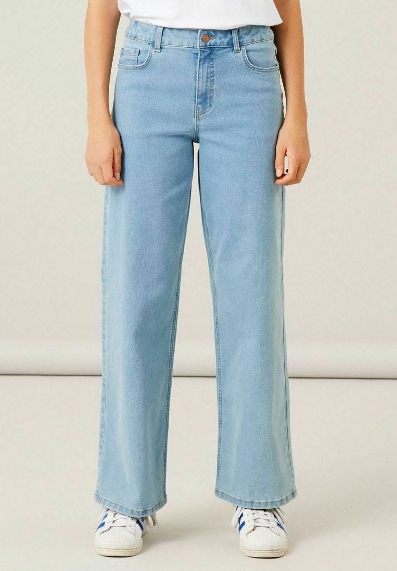 Jean large en denim bleu clair, avec une taille haute, des coutures discrètes et des poches latérales. Associé à des baskets blanches.