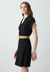 Abito camicia nero con maniche corte, bottoni frontali e una cintura testurizzata di color marrone chiaro. Tessuto morbido e orlo a ginocchio.