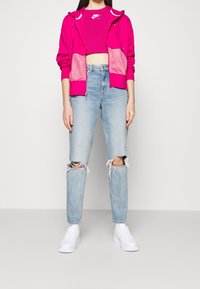 Světle růžová zipová mikina s síťovými detaily, nošená přes odpovídající crop top. Světle modré roztrhané džíny a bílé tenisky doplňují celý outfit.
