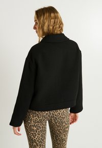 Veste noire courte avec un large col et des manches longues, fabriquée en matériau épais. Portée avec un pantalon à imprimé léopard.