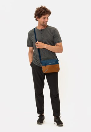 Borsa a tracolla blu e marrone con tracolla regolabile, tasche con zip e texture morbida, indossata da una persona con una maglietta grigia e pantaloni neri.