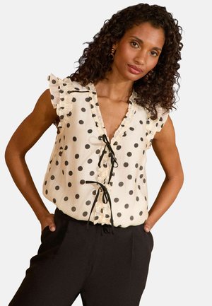 Blusa a pois in tessuto crema con maniche a ruffle, scollatura a V profonda, legami frontali e dettagli neri. Indossata con pantaloni neri.