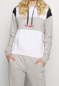 Blondýna ve šedé a bílé mikině FILA Sport a šedých teplácích, s rukama v kapsách, stojící na neutrálním pozadí.