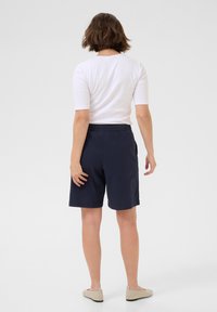 Marineblaue Shorts mit elastischem Bund, kombiniert mit einem figurbetonten weißen Oberteil. Modell von hinten gesehen, mit kurzen Ärmeln und flachen beigen Schuhen.