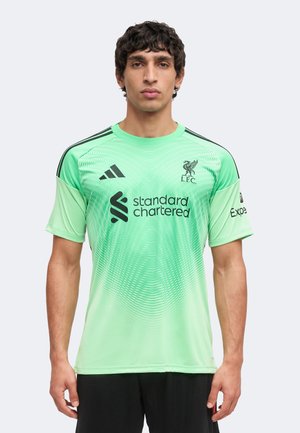 Mann trägt mintgrünes Liverpool-Fußballtrikot mit schwarzen Adidas- und Standard Chartered-Logos, steht vor einem schlichten Hintergrund.