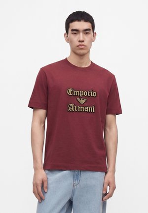 Νέος άνδρας που φοράει ένα μπορντό T-shirt Emporio Armani και ανοιχτό μπλε τζιν, στέκεται μπροστά σε μια απλή λευκή φόντο.