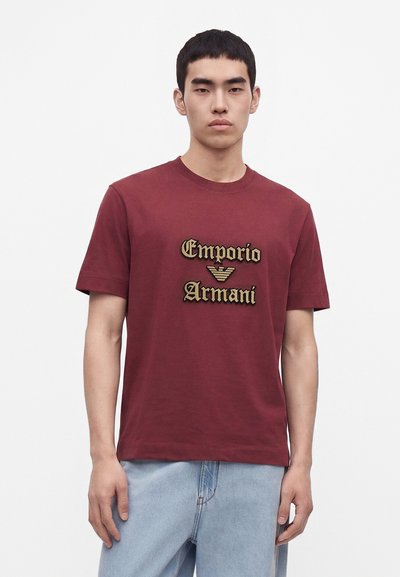 Млад мъж, носещ бордо тениска Emporio Armani и светлосини дънки, стоящ срещу бял фон.