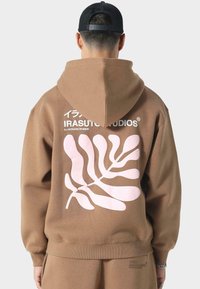 Brauner Kapuzenpullover mit einem großen pinken Blattmuster auf der Rückseite, mit Logo-Text in Weiß über der Grafik. Weicher, strukturierter Stoff.