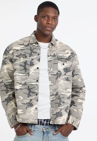 Camo-jack in grijs en beige tinten, gemaakt van lichtgewicht stof, met twee voorzakken, knoopsluiting en geborduurde tekst.