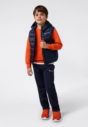 Gilet imbottito blu navy sopra un felpa arancione, abbinato a pantaloni della tuta blu navy e scarpe da ginnastica arancioni, con un logo marchiato sul petto.