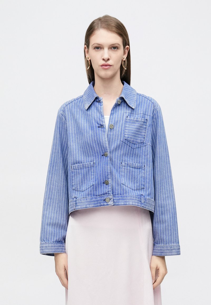 Κοντή τζιν ζακέτα σε ανοιχτό μπλε με λευκές κάθετες ρίγες, με δύο μπροστινές τσέπες, κλείσιμο με κουμπιά και γιακά με αιχμηρή σχεδίαση.