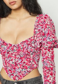 Top de corpete floral com um fundo rosa, flores brancas e roxas, mangas bufantes texturizadas e decote coração.