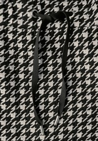 Schwarz-weißer Hahnentrittmusterstoff mit strukturierter Oberfläche und schwarzen Kordeln mit Metallspitzen zur Anpassung.