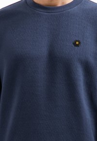 Marineblauwe sweatshirt met een gestructureerd geometrisch patroon, voorzien van een hexagonale zwart-geel badge op de linkerkant van de borst.