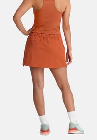 Oranje sportrok met geplooide tailleband en ritszakken. Gepaard met een aansluitend tanktop en lichtblauwe sneakers met koraalkleurige accenten.