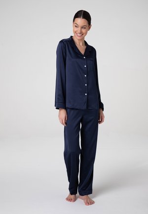 Donna in piedi a piedi nudi che indossa una camicia da pigiama in raso blu navy a maniche lunghe con bottoni e pantaloni abbinati, sorridente e che guarda in basso.