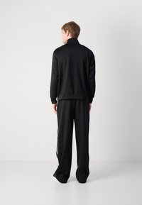 Zwart trainingspak met een ritssluiting-jack en loszittende broek. Kenmerken zijn onder andere een staande kraag en contrasterende zijstrepen op de broek.