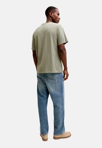 Άνδρας που φοράει ανοιχτό πράσινο μπλουζάκι, μπλε τζιν, μπεζ παπούτσια, στέκεται και κοιτάζει στο πλάι σε απλό ανοιχτόχρωμο φόντο.