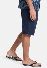 Heren navy shorts van katoen, met een platte voorkant en zijzakken. G gedragen met zwarte flip-flops. Witte achtergrond.