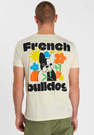 Uomo che indossa una maglietta color crema con un bulldog francese colorato in stile cartone animato, fiori e la scritta "French bulldog" sulla schiena.