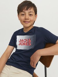 Námořnické bavlněné tričko s grafickým designem s bílými a červenými pruhy a textem "JACK & JONES". Model sedící na dřevěné židli.