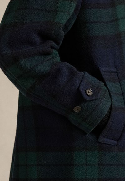 Villasegu segukaga mantel, millel on tume roheline ja tumesinine ruuduline muster. Märkimisväärsete detailide hulka kuuluvad nööbitav mansett ja tekstuuriga viimistlus.