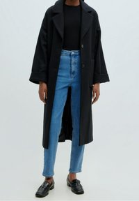 Persona vestida con un abrigo largo negro, una blusa negra, unos vaqueros de talle alto azules y unos mocasines negros, de pie contra un fondo claro y liso.