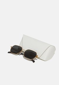 Quadratische Sonnenbrillen mit schwarzen Gläsern, goldfarbigem Metallrahmen und schwarzen Bügeln, die auf einem strukturierten weißen Etui liegen, auf dem "MICHAEL KORS" eingeprägt ist.