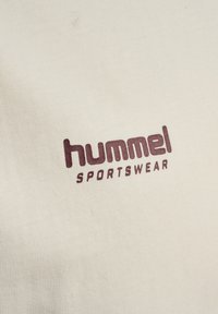 Beige sportstøjsstof med vinrødt "hummel"-logo og teksten "SPORTSWEAR"; glat tekstur med et ribbet mønster.