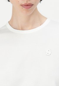 Weißes Kurzarm-T-Shirt mit Rundhalsausschnitt und einem strukturierten weißen Buchstaben "B" Emblem auf der linken Brust. Glattes Material, minimalistisches Design.