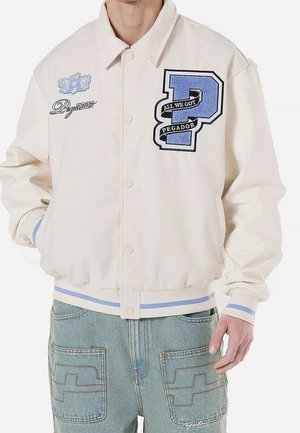 Homme portant une veste varsity crème avec des écussons bleus et noirs "P" et des patches texte, assortie à un jean denim bleu clair avec poches avant.